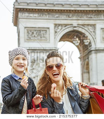 Stylish autumn in Paris. Portrait of smiling trendy mother and daughter with shopping bags near Arc de Triomphe in Paris France showing macaroons