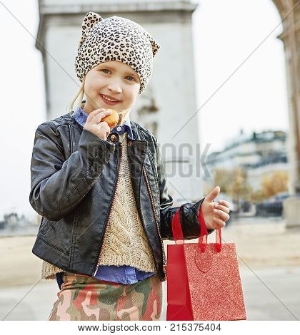 Stylish autumn in Paris. modern girl with red present bag near Arc de Triomphe in Paris holding French macaroon