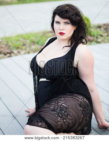 Young beautiful busty curvy plus size model with big breast in black bra and lace cape xxl woman professional makeup and hairstyle sitting outdoors