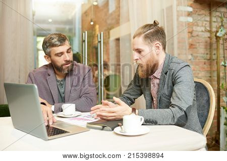 Two bearded managers analyzing results of accomplished work while sitting at restaurant with stylish interior and enjoying fragrant coffee