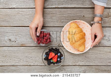 Top view on female hands serving table for breakfast. Golden crusty croissant on vintage palte and fresh organic sweet berries in glass bowls on rustic table background, copy space