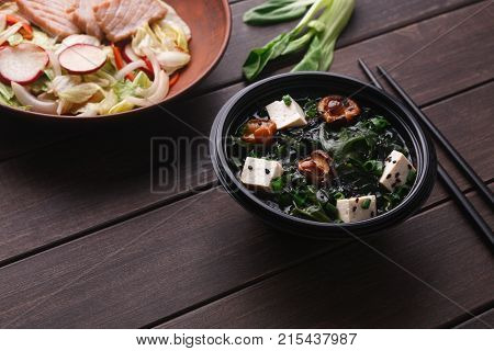 Japanese food delivery. Miso soup with tofu cheese, mushrooms and seaweed in black take away plate with fresh salad on wooden table