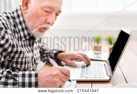 Mature man using laptop and writing in notepad at home desk. Education, recipe concept