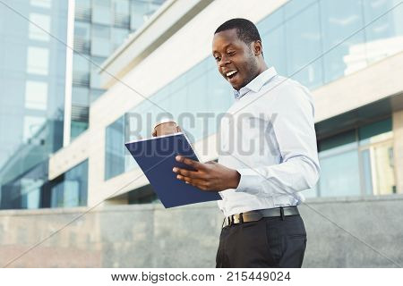 Great news. Happy successful black businessman with papers and coffee. Young executive seeing great income in the report. African-american enterpreneur outdoors near modern glass office center