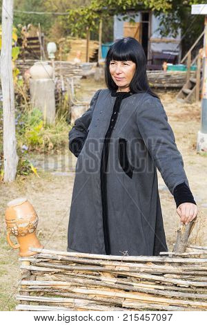 Smiling woman with black hair in ancient national clothes stands near the fence against the backdrop of rural farmstead. Ukrainian village.