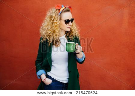 Portrait Of Pretty Woman With Curly Hair Wearing Headband, Stylish Sunglasses And Green Jacket Holdi