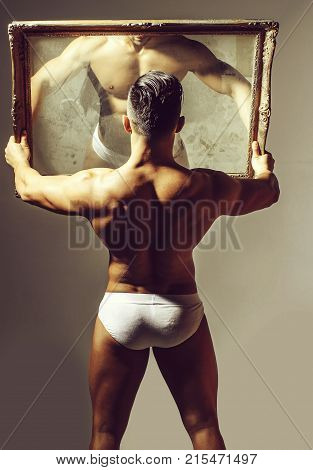 Handsome man or muscular bodybuilder macho with sexy muscle torso body with six packs and abs triceps biceps in white sexi underpants looks at mirror on grey background