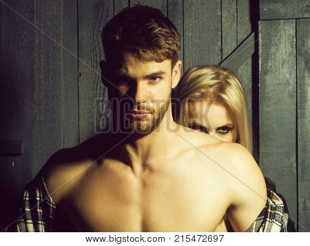 Young sexual couple of handsome serious man in checkered shirt with bare muscular chest standing before pretty blonde woman embracing and touching breast in studio horizontal picture