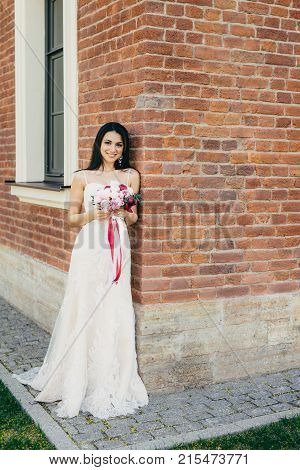 Smiling Brunette Female In Beautiful Long White Wedding Dress, Holds Bouquete, Stands Near Brick Bil
