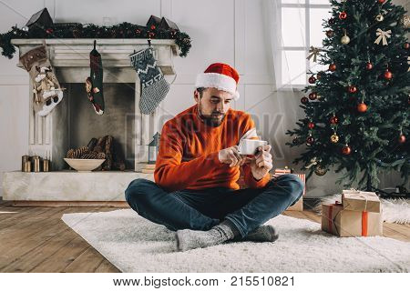 A picture of a man sitting on the white furry carpet with his legs crossed and chatting with someone on the phone. He is relaxed and peaceful. Guy is waiting for his friends to come for Christmas dinner.