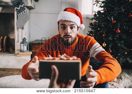 Amazed young man is sitting in a beautiful white room decorated with Christmas toys and glitter. He is getting a gift from his friend. This guy didn't expect to get it. Close up. Cut view.