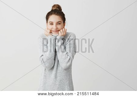 Happy fashionable caucasian girl being satisfied and glad. Female student resting at home after classes wrapping herself up in collar of grey sweater. Happiness and pleasure concept