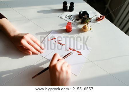 Female master of calligraphy and lettering creates red ink and pen fountain with wonderful love valentines with text I love you on day of all lovers on light table with accessories for lettering, small jars with colorful mascara and decorations in art stu