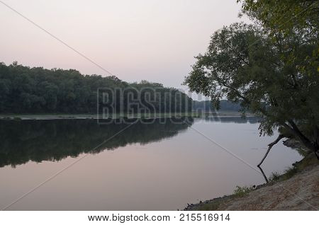 Wide river flowing across green forest. Fall. Evening. Reflections of trees in the calm water. Sundown