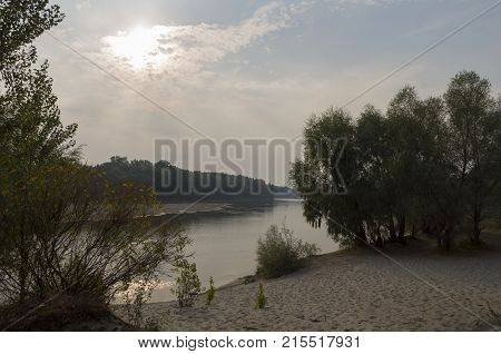Wide river flowing across green forest. Fall. Evening. Reflections of trees in the calm water. Sundown