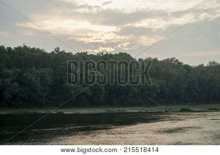 Wide river flowing across green forest. Fall. Evening. Reflections of trees in the calm water. Sundown