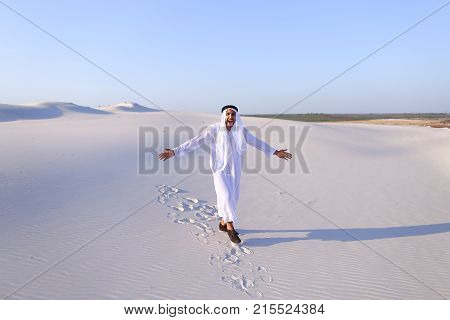 Stately young sheikh walks on foot along bottomless desert leaving traces on sand, enjoying warmth of day. Attractive Muslim smiles widely and spreads hands to sides, straightens clothes in wind and whirls around, delighted by breadth of desert. Swarthy, 