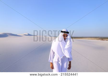 Cheerful Muslim guy walks along white sand of wide desert on warm summer evening. Arab young man in national clothes dances on spot and laughs giggly, adjusting hands with harness in wind. Swarthy, handsome Muslim with short dark hair dressed in kandura, 