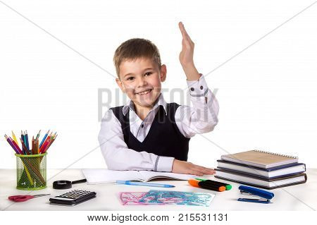 Smiling cheerful excellent pupil with hand up sitting at the table on the white background.