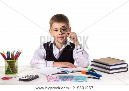 Smart serious pupil at the desk holding magnifier near the eye on the clean white background.
