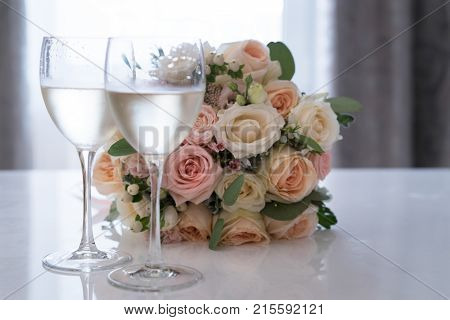 Beautiful bridal flowers bouquet on white reflecting table beside two glasses of white wine