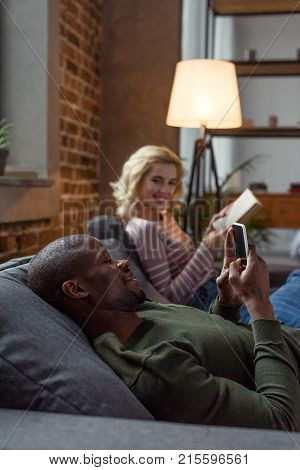 selective focus of african american man using smartphone while caucasian girlfriend reading book on sofa at home