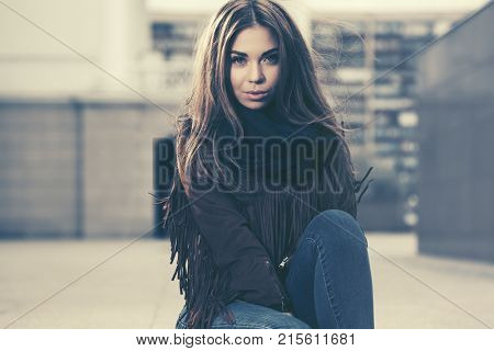 Happy young woman sitting on the sidewalk in city street. Stylish fashion model in a fringe leather suede jacket and blue jeans