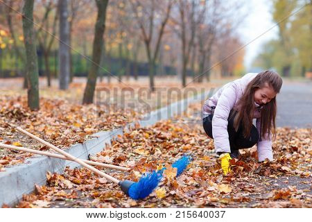 A girl in a pink jacket, sitting in an autumn park, collects the fallen leaves.