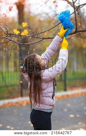 A girl in yellow, rubber gloves, reaches for a branch on a tree to take off the package, which is stuck there.