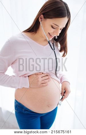 Noise in tummy.  Top view of  good looking nice pregnant woman holding stethoscope  while putting her hand on the tummy and gazing down