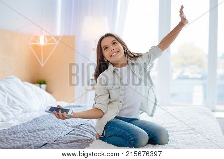 Moments with music. Fascinated happy cheerful girl sitting on the bed looking up while stretching her hands