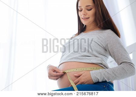 Enjoy motherhood.  Low angle of beautiful joyful pregnant woman who using meter  when  ties it around tummy and sitting