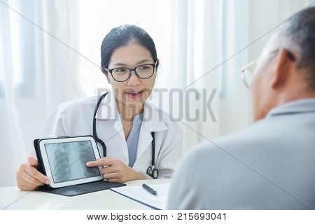 Young Asian Female Doctor wear glasses smiling and discussing with senior man patient about a chest radiograph in digital tablet computer in medical office. X-ray.