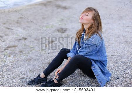 Dreamy girl relaxes and enjoys rest on seaside, throws head back and shakes hair, sitting on sandy beach on Black Sea shore on clear evening. Young European-looking woman with blond mid-length hair dressed in white jacket over which blue denim jacket, bla