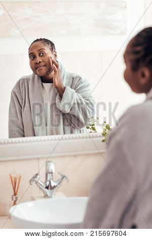 Smiling young African woman looking at her complexion in her bathroom mirror while wearing a robe in the morning
