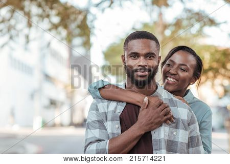 Smiling young African woman embracing her husband from behind while standing outside together on a sunny day