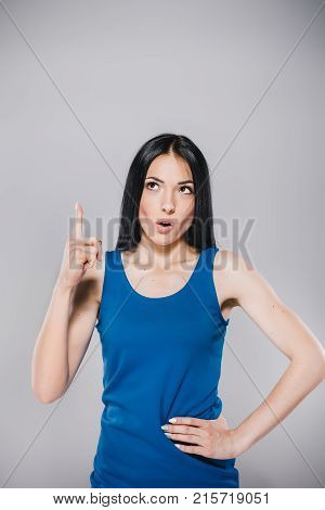 young thoughtfull teenager with long brunette hair in blue dress holding finger up in photostudio on gray background