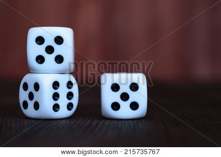 Pile of three white plastic dices on brown wooden board background. Six sides cube with black dots. Numbers 5 6.
