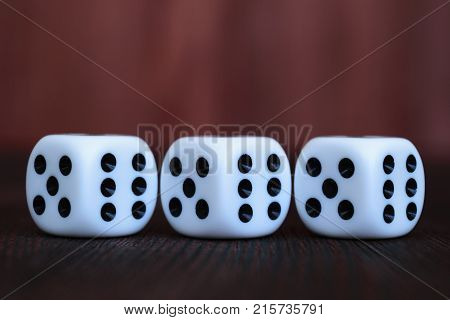 Row of three white plastic dices on brown wooden board background. Six sides cube with black dots. Numbers 5 6.
