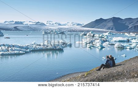 couple in warm jackets sits in Ice Lagoon in Iceland with background of glacier and sky.