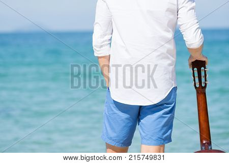 Back view of man holding guitar and standing on the beach with blue sea and blue sky bacjground. Travel relax vacation concept.