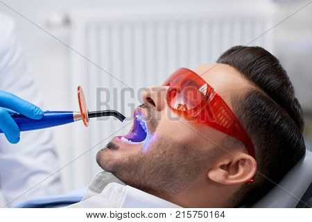 Profile close up shot of a handsome bearded man wearing protective glasses getting teeth filling done at his dental clinic copyspace technology ultraviolet UV lamp modern medicine health.