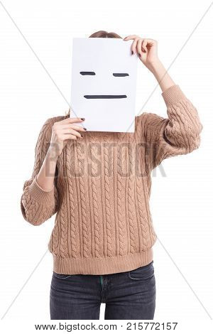 A girl in a beige sweater and jeans covers her face with a smiley face sheet with a poker face on a white isolated background