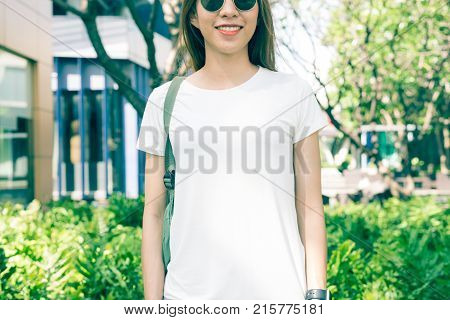 Asian hipster girl long brown hair in white blank t-shirt is standing in the middle of street. A female in street wear is standing on a green urban background. Empty mock up space for text or design.