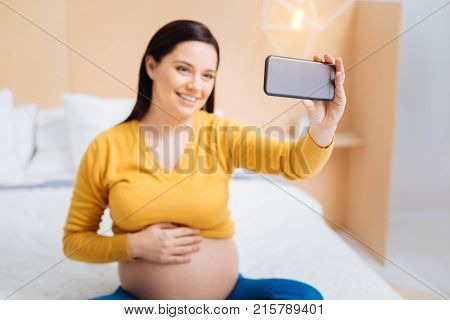 Time for selfie. Beautiful pregnancy keeping smile on her face and touching tummy while looking at camera