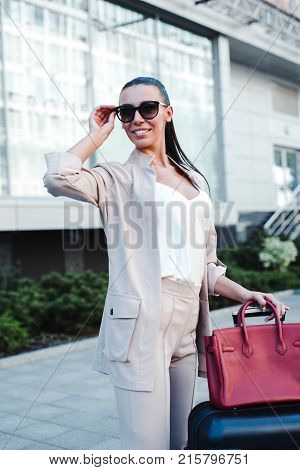 Beautiful young woman adjusting her sunglasses and looking away with smile while pulling her luggage
outdoors.