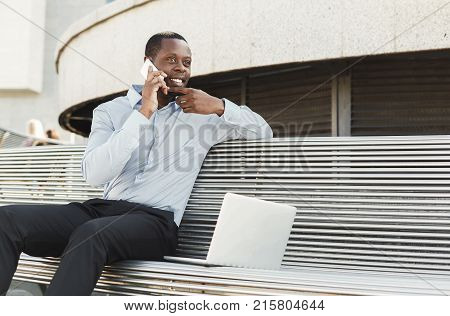 Happy black businessman working on laptop. African-american insuranse agent checking email on computer, talking on mobile, sitting on the bench outdoors near modern office center