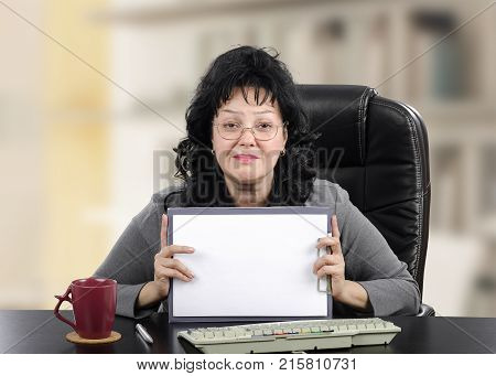 Telemental counselor holds blank page. Black-haired mature woman sits at black desk looking at the camera. Indoors office background
