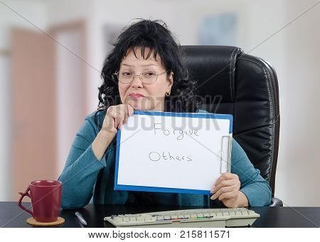 Psychologist is engaged in the use of distance counseling. Black-haired mature woman in glasses holds written message Forgive Others looking at the camera.  