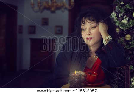 Mature woman is upset and frustrated about holiday. It's a hard time of Christmas to spend too much time alone. Black-haired woman drinks coffee head is propped up with her hand. Dark blurred indoors background